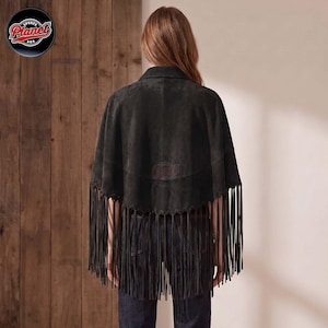 Women's genuine suede leather fringe western boho cape poncho — A dark charcoal gray suede cape featuring a scalloped hemline adorned with long, thin fringe detailing that extends from the back. The garment is worn over dark blue denim jeans, showcas