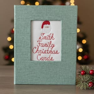 May include: A rectangular, light green fabric-covered box with a rectangular opening. The opening reveals a white background with red embroidered text that reads "Smith Family Christmas Cards" and a Santa Claus design.
