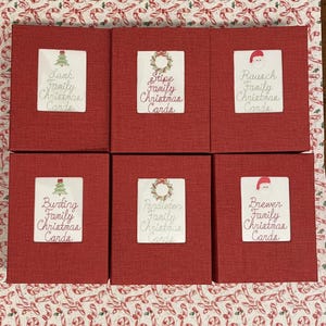May include: Six red boxes with white labels, each labeled with a family name and "Family Christmas Cards." The labels feature festive illustrations of a Christmas tree, wreath, or Santa hat. The boxes are arranged on a Christmas-themed patterned surface.