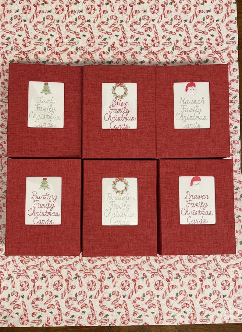 May include: Six red boxes with white labels, each labeled with a family name and "Family Christmas Cards." The labels feature festive illustrations of a Christmas tree, wreath, or Santa hat. The boxes are arranged on a Christmas-themed patterned surface.