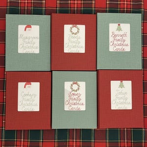 May include: Six Christmas card holders in red and green. Each card holder has a white label with a family name and "Family Christmas Cards" printed on it. The labels feature festive graphics like a Santa hat, wreath, and Christmas tree.