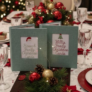 May include: Two teal fabric Christmas card holders with framed inserts. One has a Santa hat graphic and the text "Rausch Family Christmas Cards." The other has a Christmas tree graphic and the text "Willis Family Christmas Cards." The holders are on a decorated table.