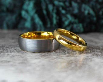 May include: Two wedding rings. One ring is gold, the other is silver with a gold interior. The rings are on a light gray surface with a dark green and gray background. The text "Two Rings One Heart" is at the bottom of the image.