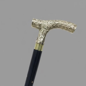 May include: A decorative walking cane with a silver-colored, ornate handle and a black shaft. The handle features detailed floral and scrollwork patterns. The cane has a brass collar where the handle meets the shaft.