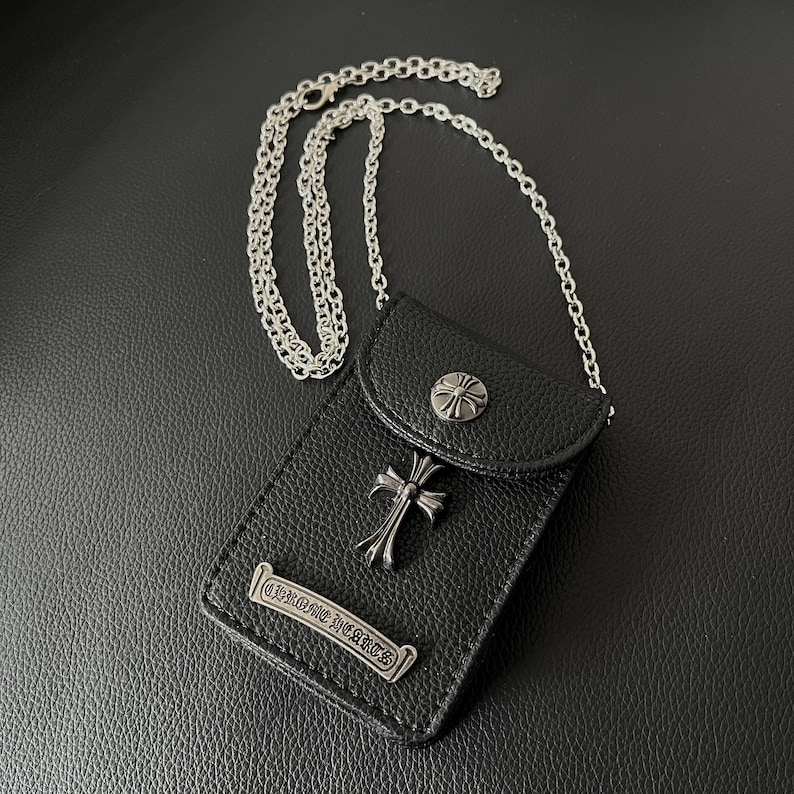May include: Black leather crossbody bag with a silver chain strap. The bag features a silver cross emblem and a decorative silver emblem with the text "CHROME HEARTS". The bag has a flap closure with a silver button.