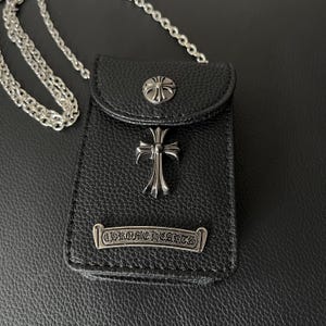 May include: Black leather crossbody bag with silver crosses and floral details. The bag has a silver chain strap and a metal plate with the text "CHROME HEARTS". The bag has a textured surface.