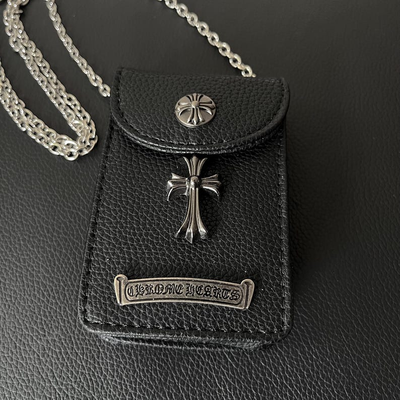 May include: Black leather crossbody bag with silver crosses and floral details. The bag has a silver chain strap and a metal plate with the text "CHROME HEARTS". The bag has a textured surface.