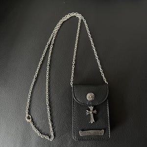 May include: Black leather phone case with a silver chain strap. The case has a silver cross and a decorative button on the flap. The bottom of the case has a silver nameplate.