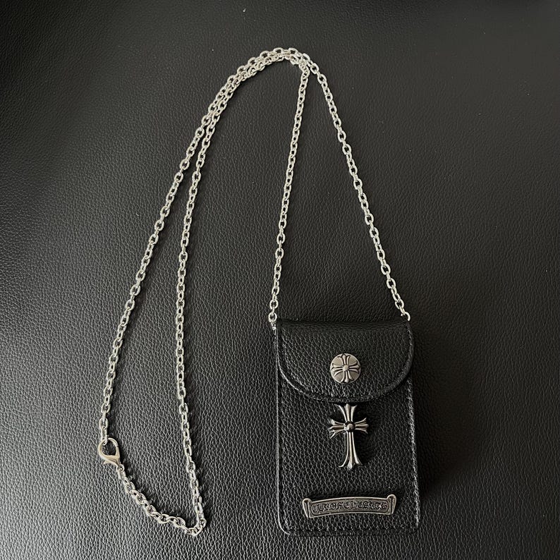 May include: Black leather phone case with a silver chain strap. The case has a silver cross and a decorative button on the flap. The bottom of the case has a silver nameplate.
