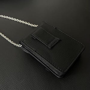 May include: A black leather wallet with a silver chain strap. The wallet has a textured surface and a flap closure. The chain is made of small, interconnected links. The wallet is designed to be worn around the neck or shoulder.