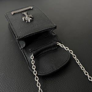 May include: Black leather pouch with a silver chain strap. The pouch has a silver cross emblem and a silver bar on the top. The pouch is open, revealing the interior. The leather has a textured appearance.