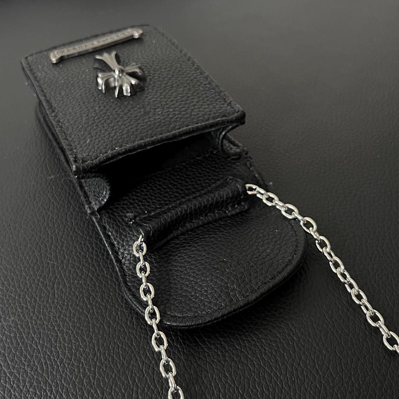 May include: Black leather pouch with a silver chain strap. The pouch has a silver cross emblem and a silver bar on the top. The pouch is open, revealing the interior. The leather has a textured appearance.