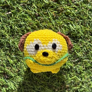 Peut inclure: Un jouet pour chien jaune crocheté à la main, avec des oreilles marron, des accents blancs autour des yeux et un nez noir. Le chien a un collier vert. Le jouet est posé sur de l'herbe verte.