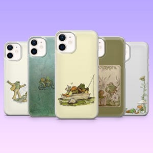 May include: Five clear smartphone cases featuring illustrations of frogs in various activities. The designs include frogs dancing, riding a tandem bicycle, fishing from a boat, interacting in a garden setting, and resting near a mushroom.