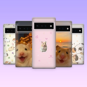 May include: A collection of phone cases with various designs. One case is light pink with a hamster illustration and hearts. Other cases feature hamster photos, a sunset, and a pattern of guinea pigs and flowers. The cases are displayed against a purple background.