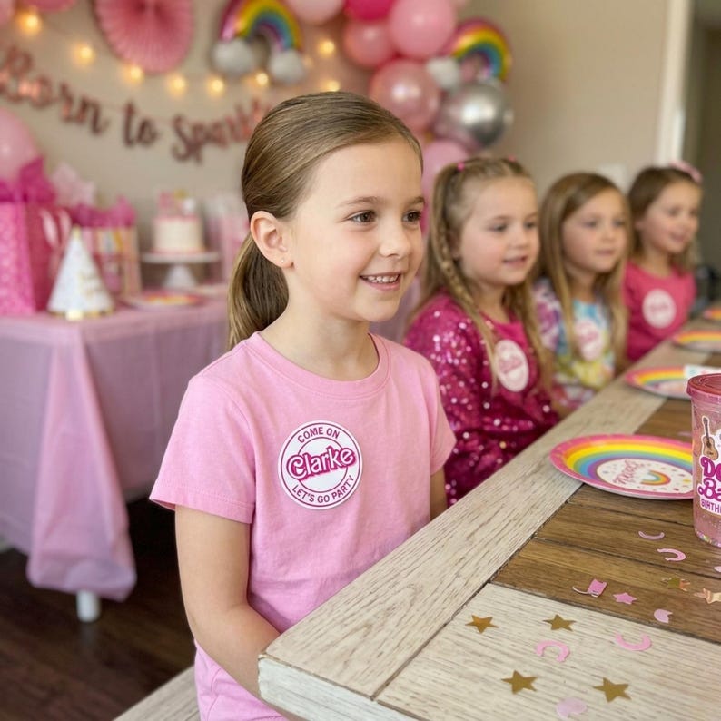 May include: A young girl wearing a pink t-shirt with the text "Come on Clarke Let's Go Party" at a birthday party. The party has pink decorations, balloons, and a table set with plates and cups.