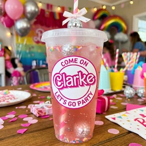 May include: A clear plastic cup filled with a pink beverage, a striped straw, and a disco ball topper. The cup has a white sticker that says "Come on Clarke Let's Go Party". The drink is decorated with sprinkles.
