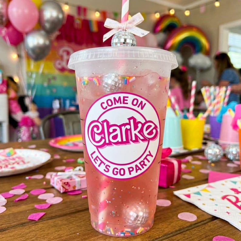 May include: A clear plastic cup filled with a pink beverage, a striped straw, and a disco ball topper. The cup has a white sticker that says "Come on Clarke Let's Go Party". The drink is decorated with sprinkles.