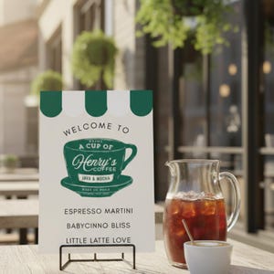 以下が含まれることがあります： 「Welcome to Henry's Coffee」と書かれた緑と白のデザインの看板。黒い金属製のスタンドに立てられています。テーブルには、アイスコーヒーの入ったピッチャーとコーヒーカップがあります。看板にはコーヒーの飲み物が記載されています。