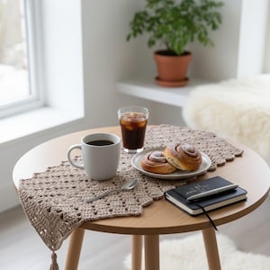 May include: A round wooden table with a crocheted runner, coffee, pastries, and a notebook. The beige runner has a scalloped edge and a tassel. A cup of coffee and a glass of iced coffee are on the table.