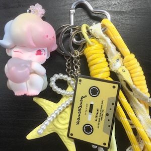 May include: A collection of keychains and charms, including a cartoon figure, a yellow starfish with a pearl strand, and a yellow cassette tape charm with the text "WanaShop". The keychains have yellow and white accents.