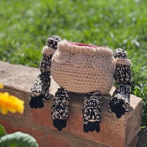 Puede incluir: Una olla beige tejida a ganchillo con cuatro patas estampadas en blanco y negro. La olla tiene un diseño en espiral y un interior rojo. El objeto está sobre una superficie de ladrillo con un fondo verde borroso.