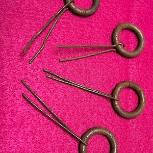 以下が含まれることがあります： 明るいピンク色の表面に、ワイヤーのピンが付いた4つのアンティークメタルリングが置かれています。リングは濃い茶色で古くなっており、ワイヤーピンは細くてわずかに曲がっています。これらのアイテムはヴィンテージまたは再利用されたもののようです。