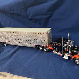 May include: A die-cast model semi-truck featuring a black cab with orange fenders and chrome accents, attached to a long silver livestock trailer. The trailer has a metallic finish with rows of ventilation slots along the side panels.