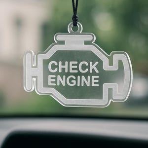 Könnte beinhalten: Klarer Auto-Lufterfrischer aus Acryl in Form eines Motors mit den Worten "CHECK ENGINE" in der Mitte. Der Lufterfrischer hängt an einer schwarzen Schnur, mit einem verschwommenen grünen Hintergrund.