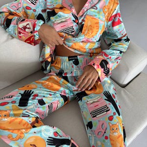 May include: Light blue pajama set with a vibrant print featuring orange cats, typewriters, guitars, and black gloves. The long-sleeved top has pink trim and a button-down front. The pajama pants have a matching print. The brand name "COCOLARE" is visible on the pants.