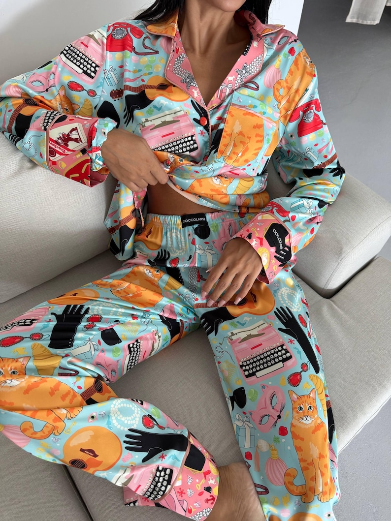 May include: Light blue pajama set with a vibrant print featuring orange cats, typewriters, guitars, and black gloves. The long-sleeved top has pink trim and a button-down front. The pajama pants have a matching print. The brand name "COCOLARE" is visible on the pants.