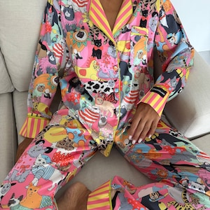 May include: A vibrant pink pajama set featuring a playful cat-themed print. The long-sleeved top and pants are adorned with various cat illustrations in a colorful, cartoonish style. Yellow and pink striped trim accents the collar, cuffs, and pant hems.