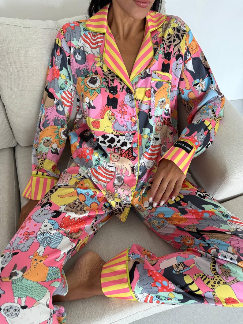 May include: A vibrant pink pajama set featuring a playful cat-themed print. The long-sleeved top and pants are adorned with various cat illustrations in a colorful, cartoonish style. Yellow and pink striped trim accents the collar, cuffs, and pant hems.