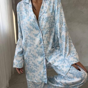 Pode incluir: Conjunto de pijama azul claro e branco com um padr&atilde;o floral e de p&aacute;ssaros. A parte superior de manga comprida tem uma frente abotoada e um bolso no peito. As cal&ccedil;as s&atilde;o compridas e soltas. Acabamento branco.