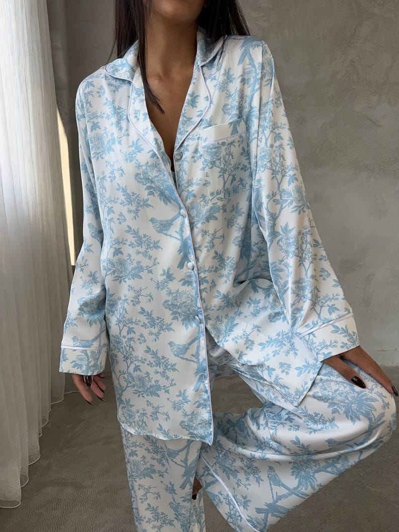 Pode incluir: Conjunto de pijama azul claro e branco com um padr&atilde;o floral e de p&aacute;ssaros. A parte superior de manga comprida tem uma frente abotoada e um bolso no peito. As cal&ccedil;as s&atilde;o compridas e soltas. Acabamento branco.
