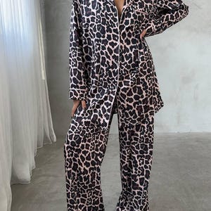 May include: A two-piece pajama set featuring a leopard print pattern in shades of brown and black on a light pink background. The set includes a button-down shirt with a collar and long sleeves, and matching wide-leg pants. White trim accents the edges.