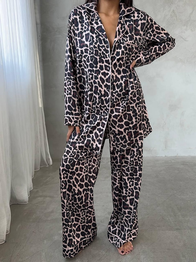 May include: A two-piece pajama set featuring a leopard print pattern in shades of brown and black on a light pink background. The set includes a button-down shirt with a collar and long sleeves, and matching wide-leg pants. White trim accents the edges.
