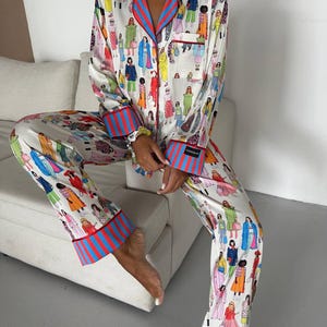 May include: White pajama set with a colorful illustration of people in various outfits. The set features red and blue striped trim on the collar, cuffs, and pant hems. The long-sleeved top has a pocket on the chest.