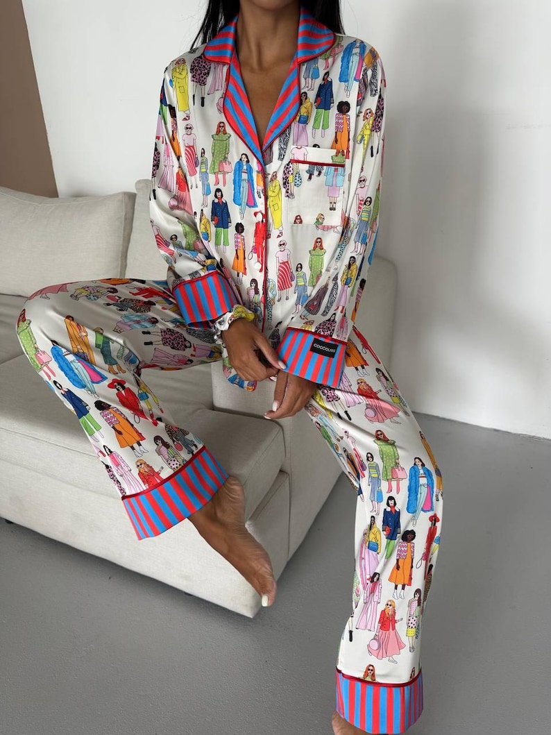 May include: White pajama set with a colorful illustration of people in various outfits. The set features red and blue striped trim on the collar, cuffs, and pant hems. The long-sleeved top has a pocket on the chest.
