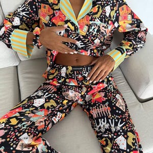 May include: Black pajama set with a colorful print featuring playing cards, leopards, and other whimsical designs. The long-sleeved top has a collared neckline and striped accents in yellow and light blue. The matching pants complete the set.