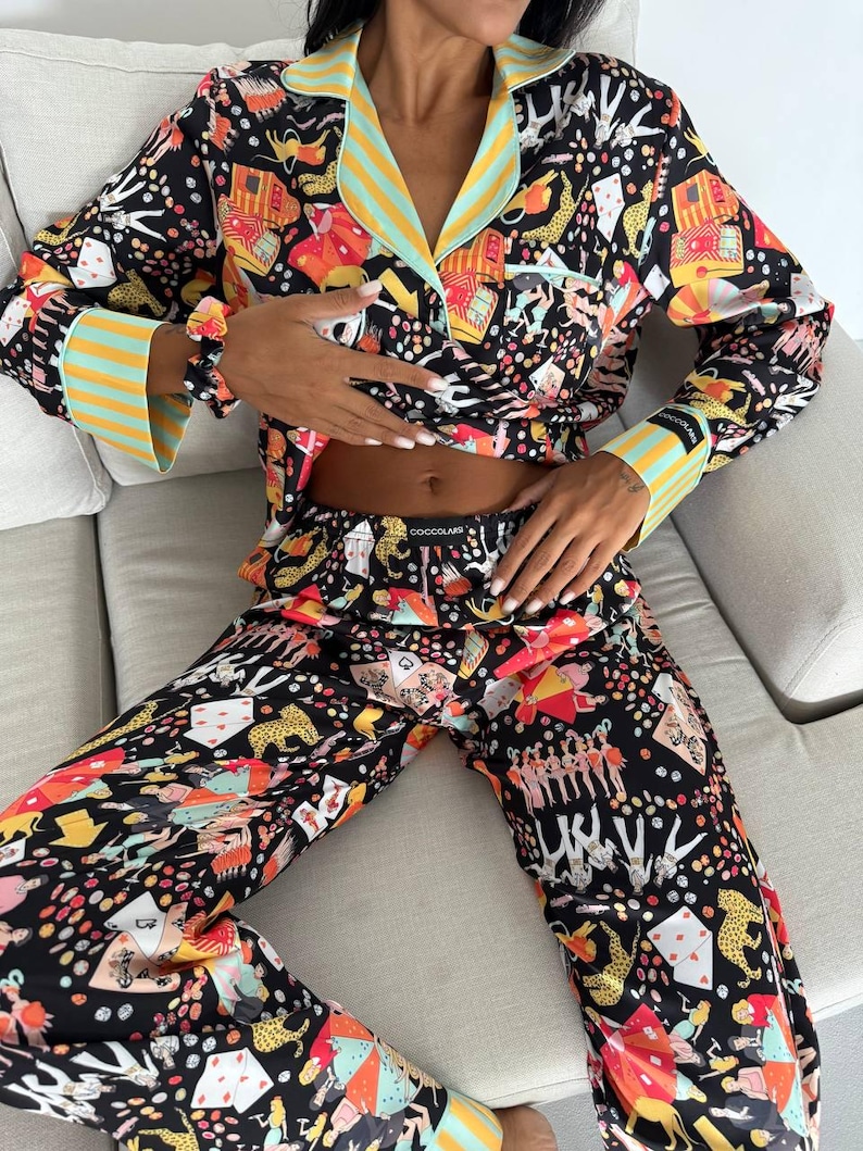 May include: Black pajama set with a colorful print featuring playing cards, leopards, and other whimsical designs. The long-sleeved top has a collared neckline and striped accents in yellow and light blue. The matching pants complete the set.