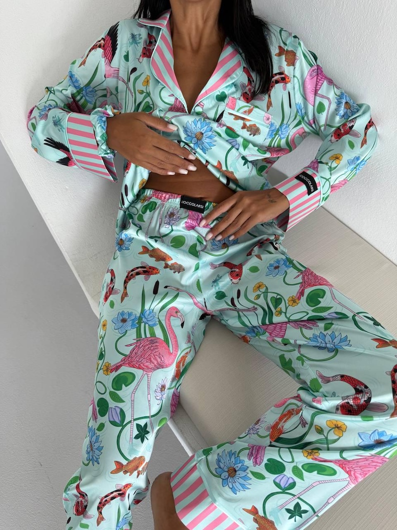 May include: Light blue pajama set with a vibrant print of koi fish, flamingos, and floral elements. The long-sleeved top and pants feature pink and white striped accents on the cuffs and collar. The pajama set is made of a silky material.