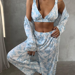 May include: A three-piece pajama set in white and light blue. The set includes a bralette, wide-leg pants, and a long-sleeved robe. The fabric features a bird and floral pattern. The bralette has adjustable straps. The pajama set is made of a soft, lightweight material.