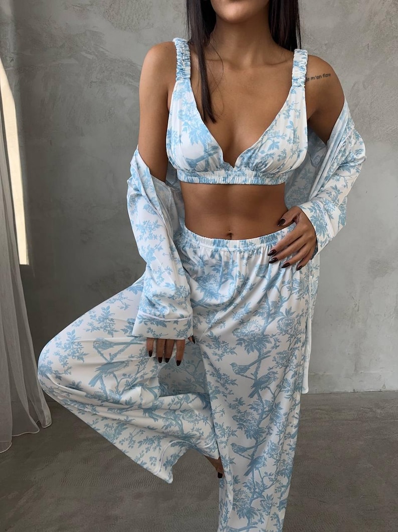 May include: A three-piece pajama set in white and light blue. The set includes a bralette, wide-leg pants, and a long-sleeved robe. The fabric features a bird and floral pattern. The bralette has adjustable straps. The pajama set is made of a soft, lightweight material.