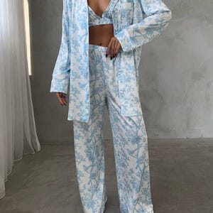 May include: A three-piece pajama set in a light blue and white floral pattern. The set includes a long-sleeved button-down shirt, a bralette, and wide-leg pants. The fabric has a silky sheen.