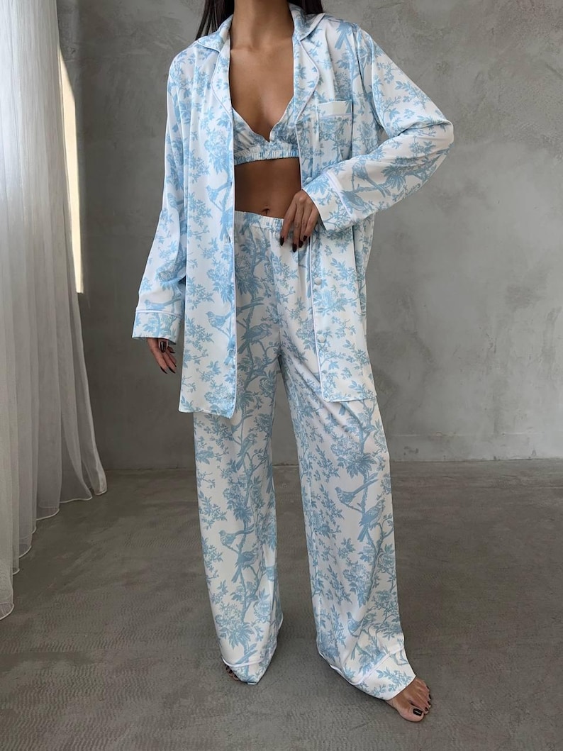 May include: A three-piece pajama set in a light blue and white floral pattern. The set includes a long-sleeved button-down shirt, a bralette, and wide-leg pants. The fabric has a silky sheen.