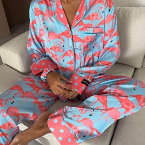 May include: Light blue pajama set with a flamingo pattern. The set includes a button-down shirt with a chest pocket and pants. The cuffs and collar are coral with blue polka dots. The pajamas are made of a silky material.