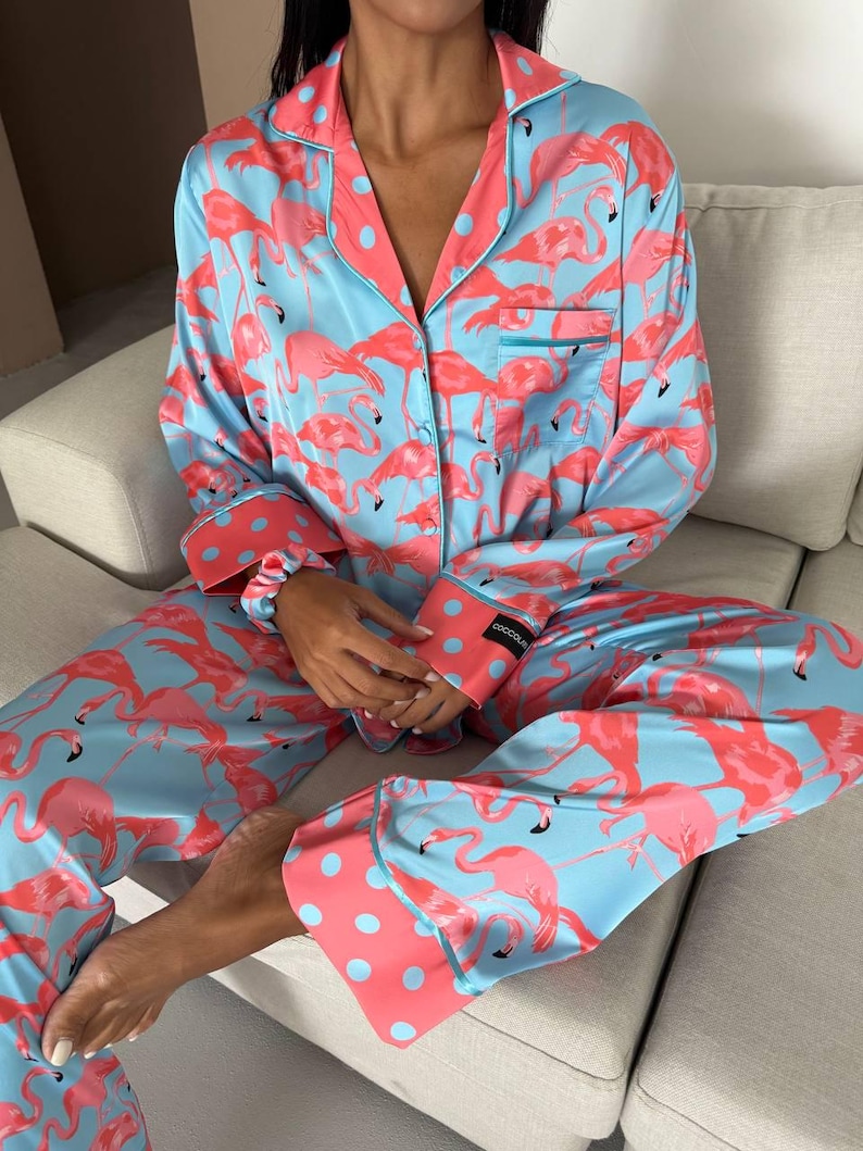 May include: Light blue pajama set with a flamingo pattern. The set includes a button-down shirt with a chest pocket and pants. The cuffs and collar are coral with blue polka dots. The pajamas are made of a silky material.