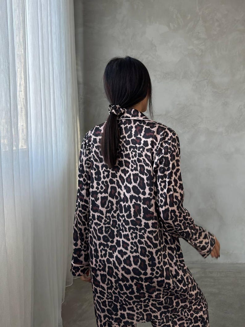 May include: A person wearing a leopard print pajama set. The pajamas are a light brown color with dark brown spots. The set has white piping around the collar and cuffs. The person has dark hair pulled back in a ponytail.
