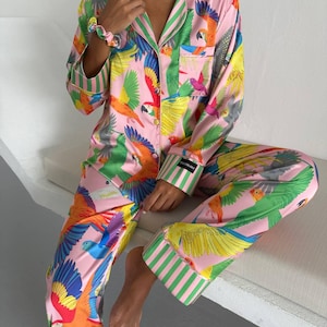 May include: Pink pajama set with a vibrant parrot print in shades of green, yellow, orange, and blue. The long-sleeved top and pants feature green and white striped accents. The set is made of a silky material.